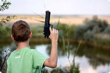 A young boy standing by a pond, holding a gun in his hands.