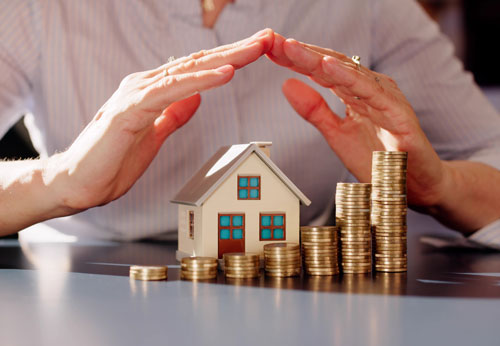 Hands protecting a model house and stacks of coins: home equity protection.
