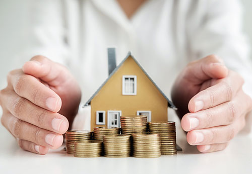 Hands protecting a model house and stacks of coins.
